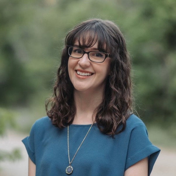 Woman with glasses and a blue shirt standing outdoors with a blurred green background