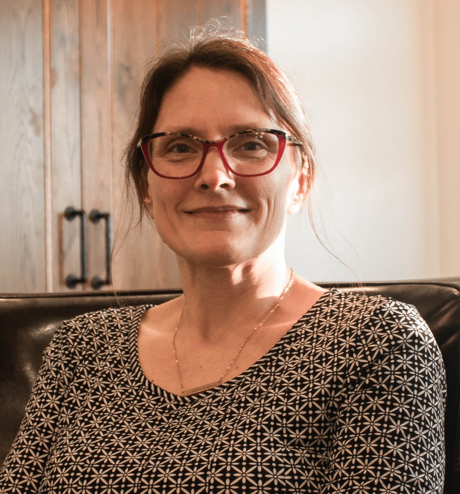 Woman wearing red glasses and a patterned shirt sitting on a couch.