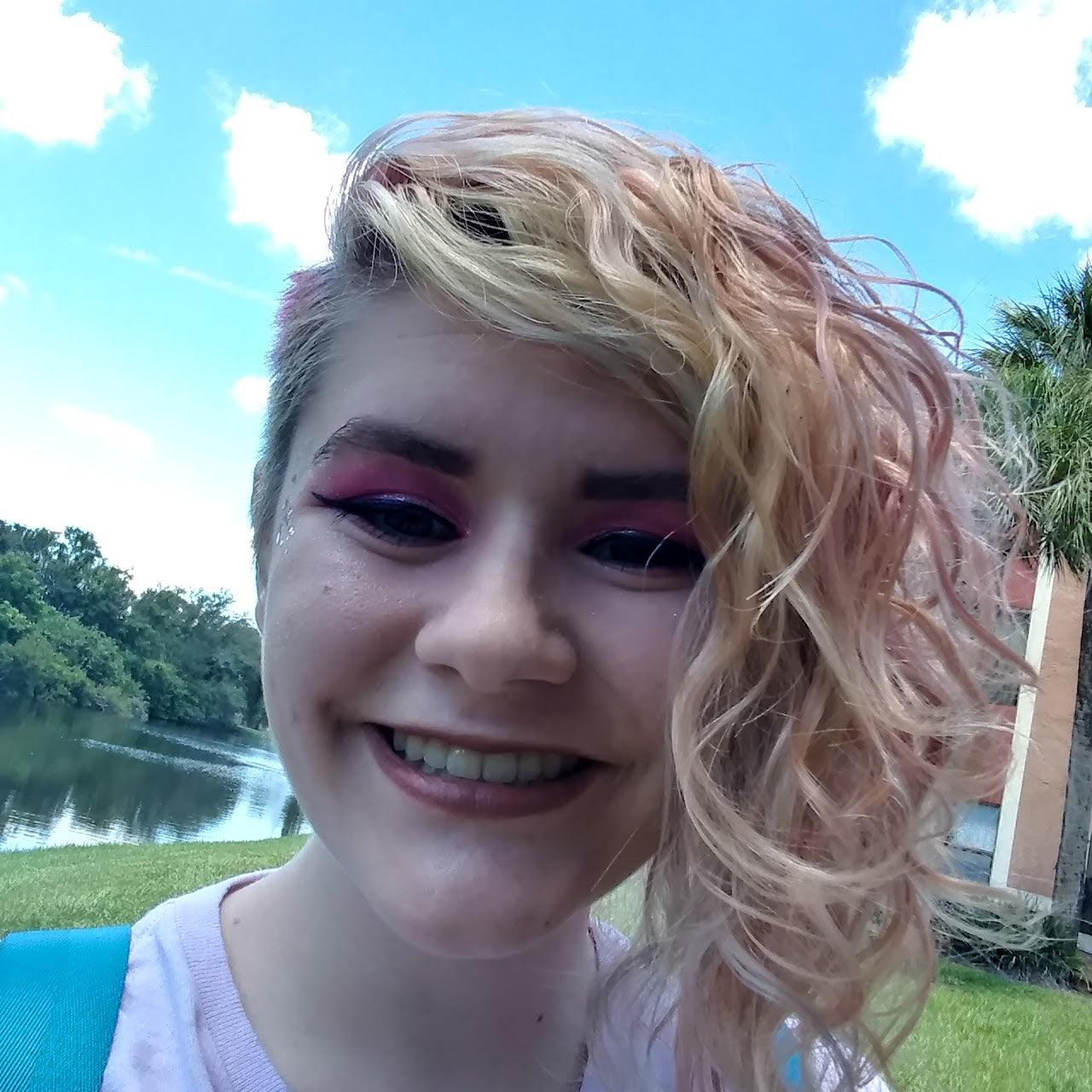 Person with pink hair and makeup smiling outdoors with a lake and trees in the background