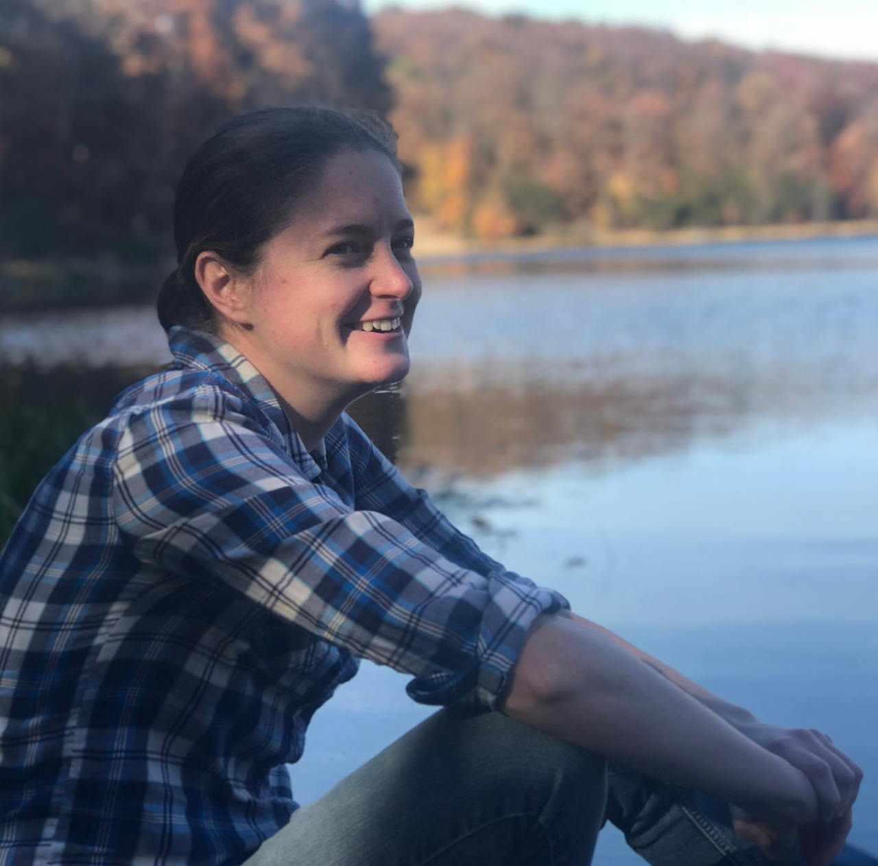 Person sitting by a lake with a plaid shirt and jeans, surrounded by trees with autumn foliage.